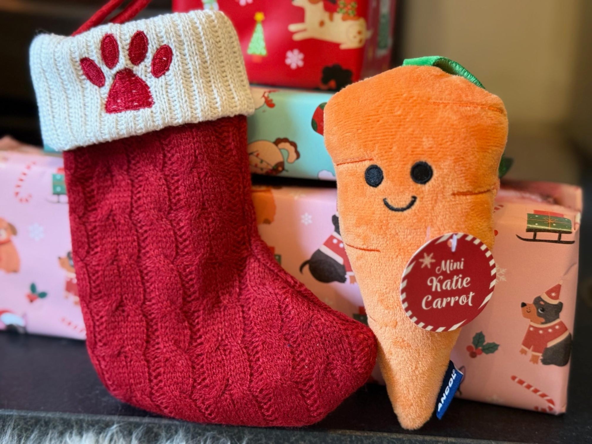 Red knitted Christmas stocking with a plush carrot toy and gift boxes in the background.