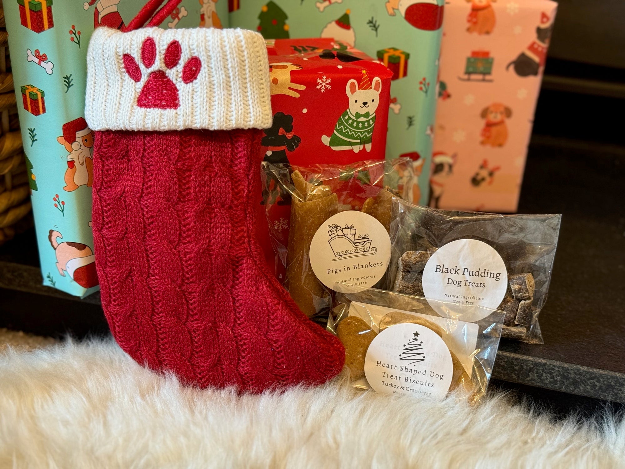 Red knitted Christmas stocking with a paw print, dog treats, and gift boxes in the background.