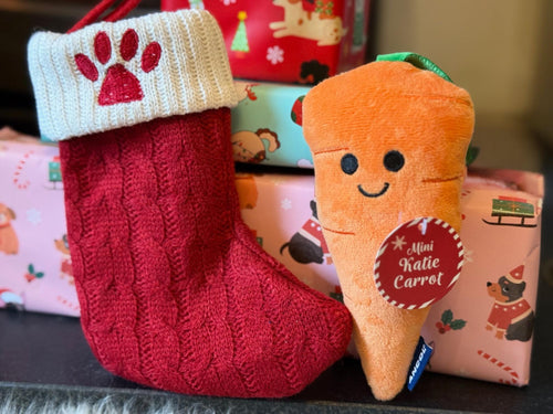 Red knitted Christmas stocking with a plush carrot toy and gift boxes in the background.