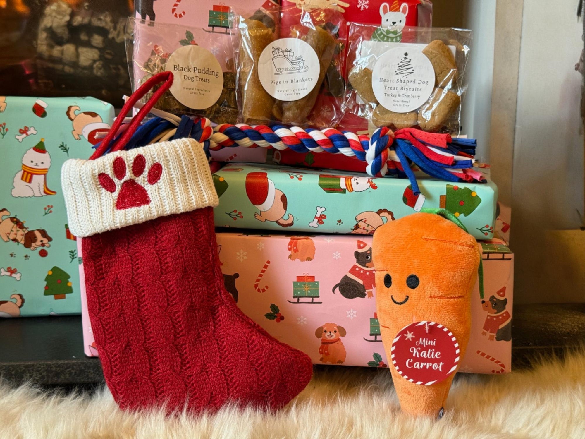 Red knitted stocking with a paw design, dog treats, and a plush carrot toy on patterned paper.
