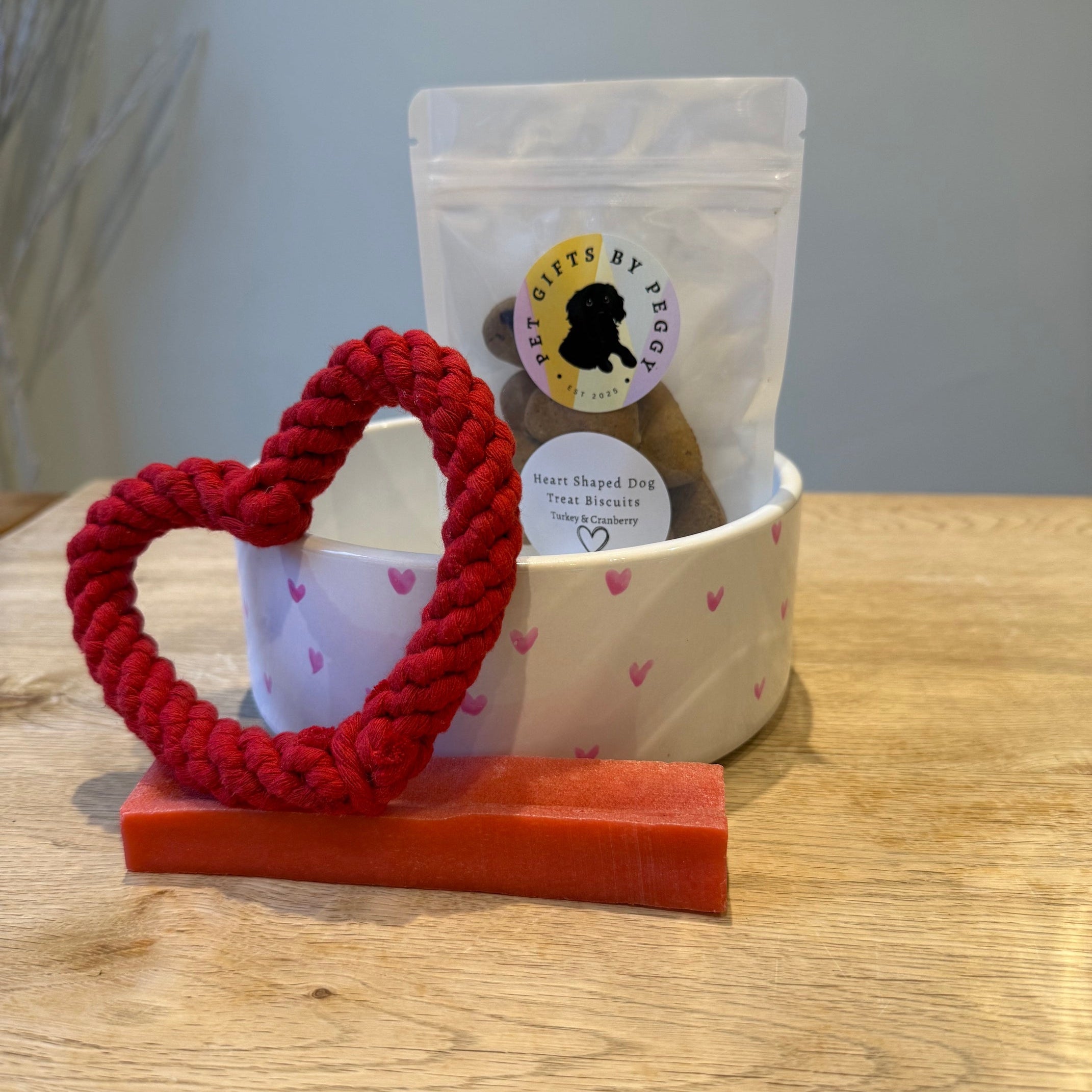 Red heart dog toy, pink yak chew dog treat and heart dog treats in packaging in a white dog bowl decorated with pink hearts on a wooden surface